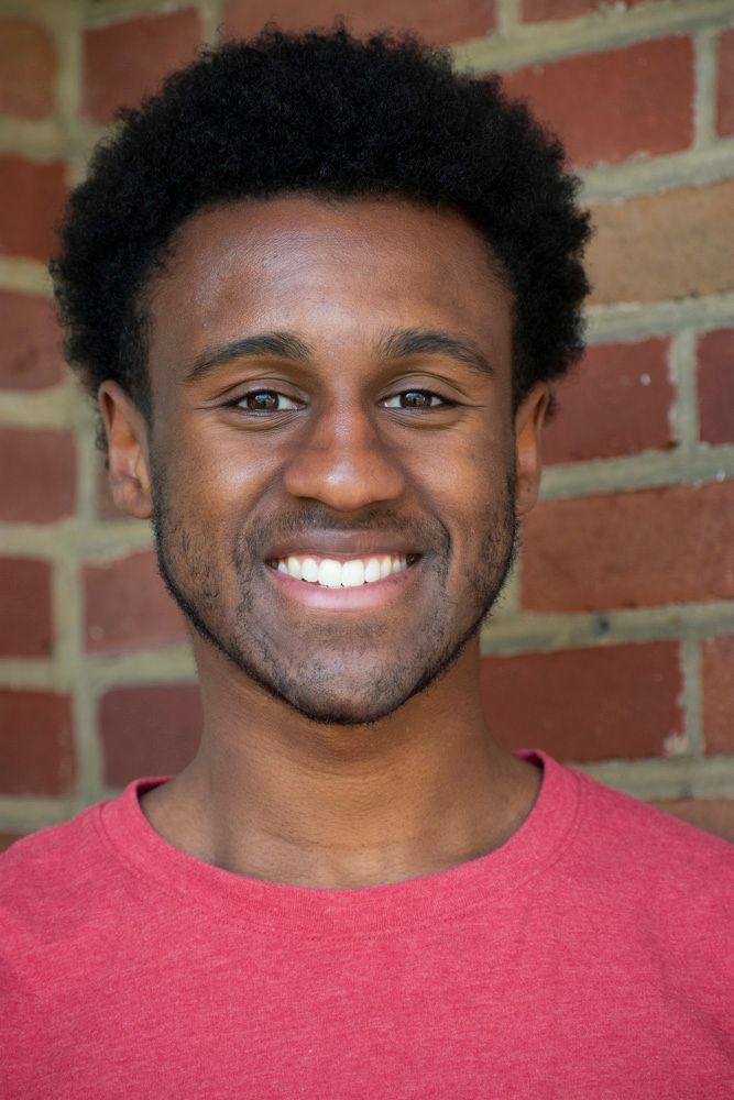 A person with dark, curly hair wearing a red shirt, smiling at the camera in front of a brick wall.