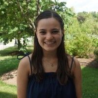 A smiling person with long dark hair wears a navy blue sundress, posing outdoors against a background of green trees.