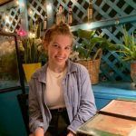 A person smiling while sitting at a table in a dimly lit space with a blue latticed wall and potted plants in the background.