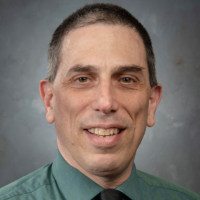 A headshot of a smiling person wearing a green shirt and a black tie, set against a plain gray background.
