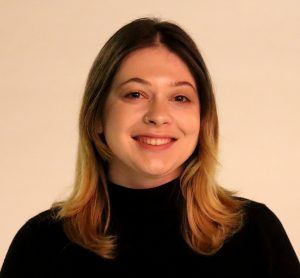 A smiling person with shoulder-length, two-toned brown and blonde hair wears a black turtleneck against a plain background.