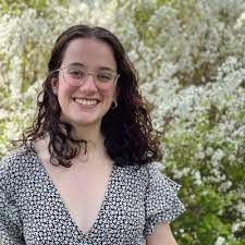 A person with dark, curly hair and glasses smiling in front of a blooming white tree.