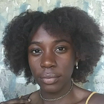 A close-up portrait of a person with dark curly hair and a gold necklace against a textured, light-colored background.