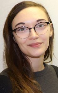Portrait of a person with long dark hair and thin-rimmed glasses, smiling slightly against a plain white background.