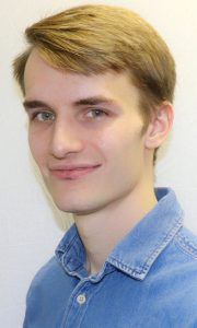 A person with short, light-brown hair, smiling softly, wearing a blue collared shirt against a plain, light background.