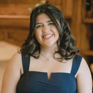 Smiling person in a sleeveless navy blue dress with a small necklace, posed indoors against a blurred wooden background.