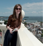 A person wearing sunglasses and a brown top leans on a white balcony railing overlooking a coastal city and the ocean.
