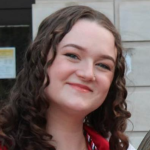 A smiling person with long, curly brown hair, wearing a red shirt, outdoors against a light-colored stone wall.