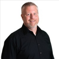 A professional headshot of a person with short hair and a beard, smiling and wearing a black collared shirt.