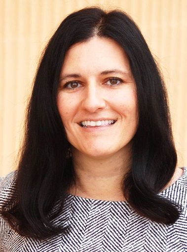 A smiling person with dark hair, wearing a patterned top, posed against a light, vertical-lined background.