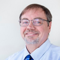 A smiling person with glasses and a goatee wearing a light blue button-down shirt and a blue patterned tie.