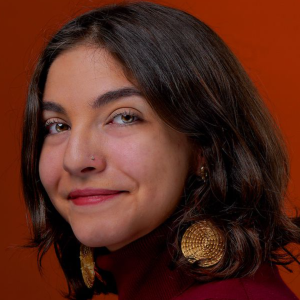 A person with shoulder-length brown hair and large gold earrings smiling against a solid, burnt-orange background.