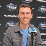 A person smiling at a podium with a microphone, set against a black backdrop with repeating March Madness logos.