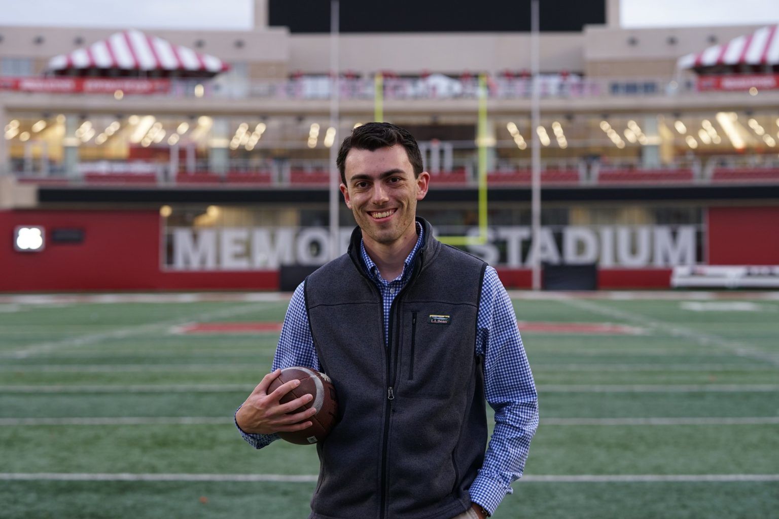 A person in a blue shirt and dark vest holds a football on a football field in front of Memorial Stadium.