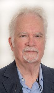 A professional headshot of a person with white hair and a goatee, wearing a blue button-up shirt and a dark blazer.