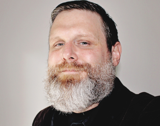 A person with a grey, full beard and short dark hair looks toward the camera with a slight smile against a light background.