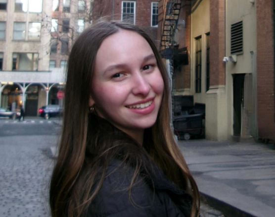 A person with long brown hair smiles over their shoulder while standing on a city street with brick buildings.