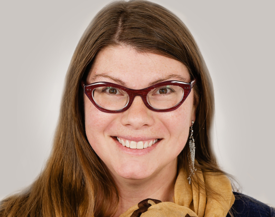 A smiling person with long brown hair, wearing maroon cat-eye glasses and a gold scarf against a neutral background.
