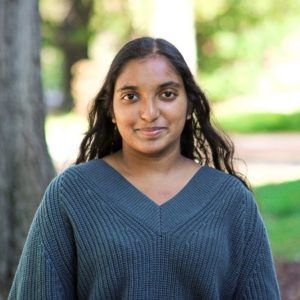 A person with long, dark, wavy hair wearing a teal V-neck sweater, posed outdoors against a blurred tree and green background.