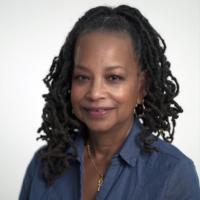A person with dark locs wearing a blue collared shirt and an ankh necklace, centered against a plain white background.