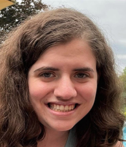 A smiling person with wavy brown hair outdoors in natural lighting.