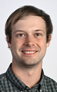 A person with short brown hair and a light smile, wearing a green plaid collared shirt against a white background.