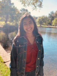 A smiling person wearing a dark denim jacket and red top, posing outdoors near a pond on a sunny day.