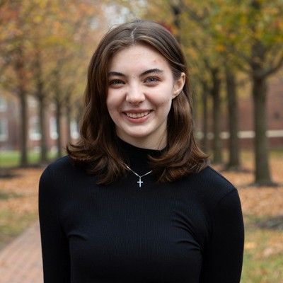 A person with shoulder-length brown hair smiles, wearing a black mock-neck top and a small cross necklace outdoors.