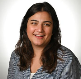 A person with long, dark hair wearing a blue and white pinstriped shirt, smiling against a plain white background.