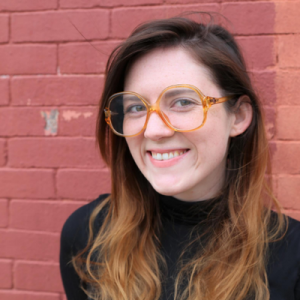 A smiling person with long brown hair wearing large, amber-tinted glasses, standing in front of a red brick wall.