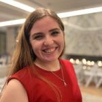 A smiling person with long light brown hair wearing a red sleeveless top and a gold pendant necklace indoors.
