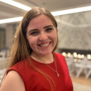 Smiling person with long hair wearing a red top and a silver necklace, set against a blurred indoor backdrop.