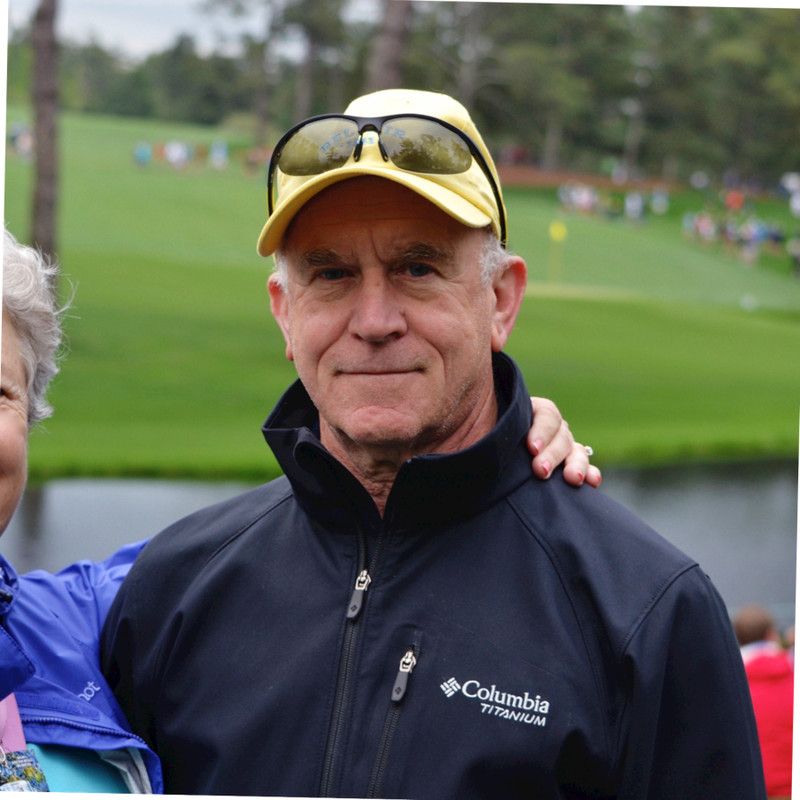 A man in a yellow cap, sunglasses, and navy Columbia jacket stands outdoors on a golf course.
