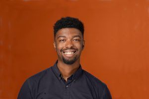 A smiling person in a dark button-down shirt centered against a plain, solid orange background.