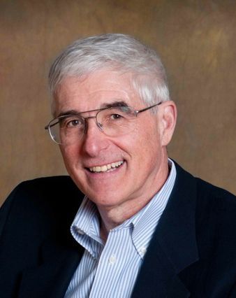 A smiling person with gray hair and glasses, wearing a striped shirt and dark blazer against a plain brown background.