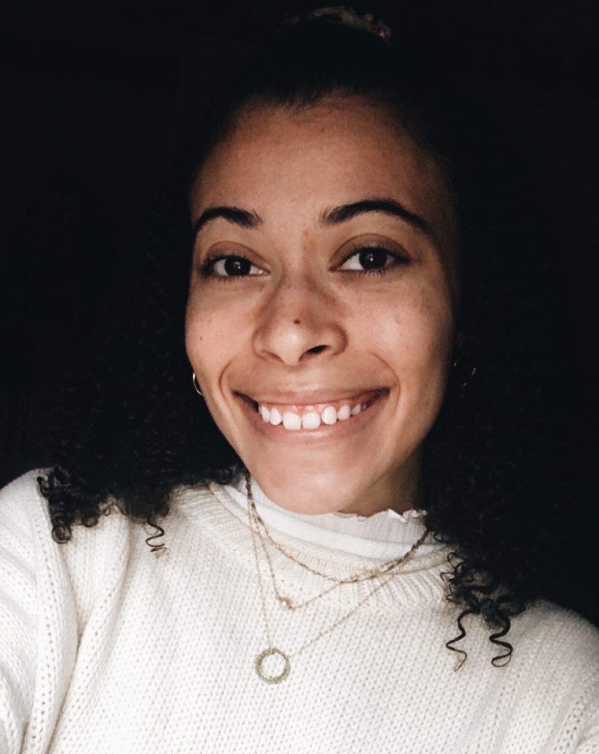 A smiling person with curly dark hair wearing a white sweater and layered necklaces against a dark background.