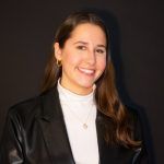 A smiling person wearing a white turtleneck and black blazer against a dark background.