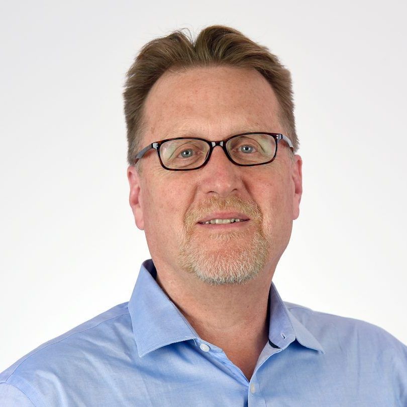 A person with short brown hair and glasses wearing a light blue button-down shirt against a white background.