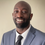 A man wearing a blue suit, white shirt, and black tie, smiling against a neutral background.