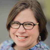 A person with shoulder-length brown hair and glasses smiling warmly toward the camera against a neutral background.