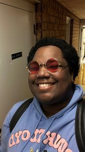 A smiling person wears red-tinted circular sunglasses and a blue hooded sweatshirt in a hallway.