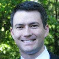 A person with dark hair and a light complexion smiling warmly, wearing a white shirt and dark jacket against greenery.