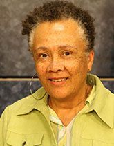 A person with short, textured hair and hoop earrings, wearing a light green collared jacket, smiling against a dark backdrop.