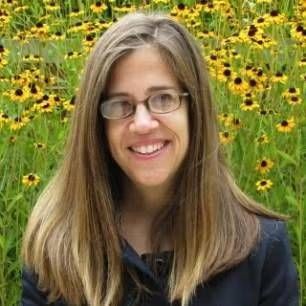 A smiling person with long light brown hair and glasses, set against a background of yellow wildflowers.