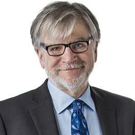 Professional headshot of a person with gray hair, glasses, and a beard, wearing a suit and a patterned blue tie.