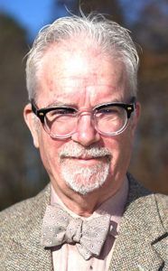 A person with short grey hair and glasses, wearing a bow tie and a textured blazer, centered against a blurred background.