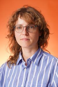 A person with wavy reddish-brown hair and glasses wearing a blue-and-white striped button-down shirt against an orange background.