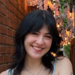 A smiling person with dark hair and bangs stands in front of a brick wall decorated with hanging floral lights.