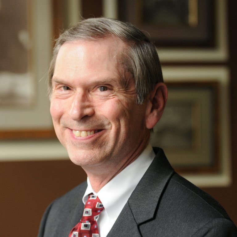 Smiling man in a grey suit and red patterned tie against a professional office background with framed art.