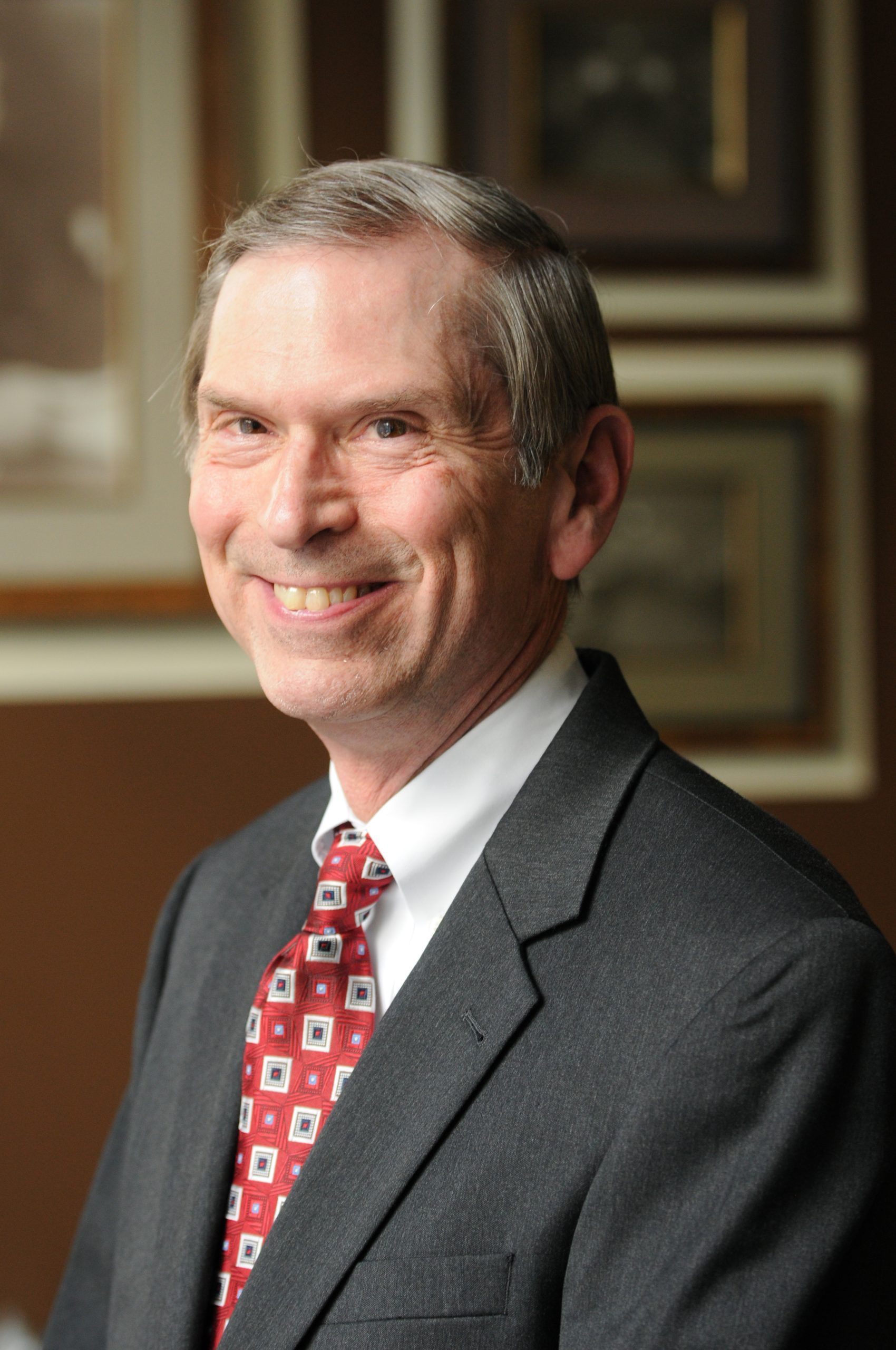 A man in a suit and patterned red tie smiling, with framed artwork on a brown wall in the background.